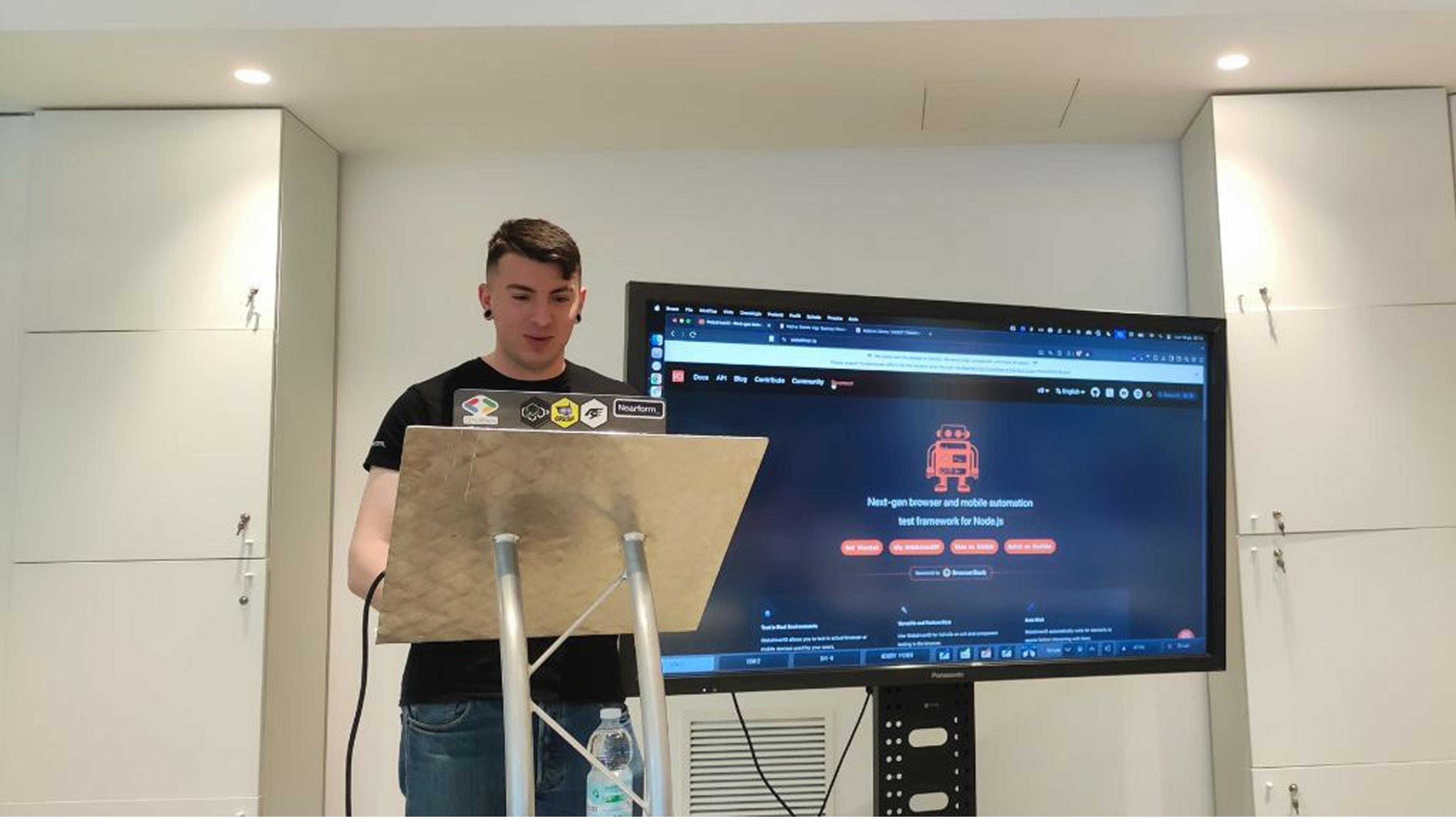 Ludovico Besana speaking as a speaker at the Ministry of Testing Milan meetup, standing behind a lectern with a projector screen in the background.