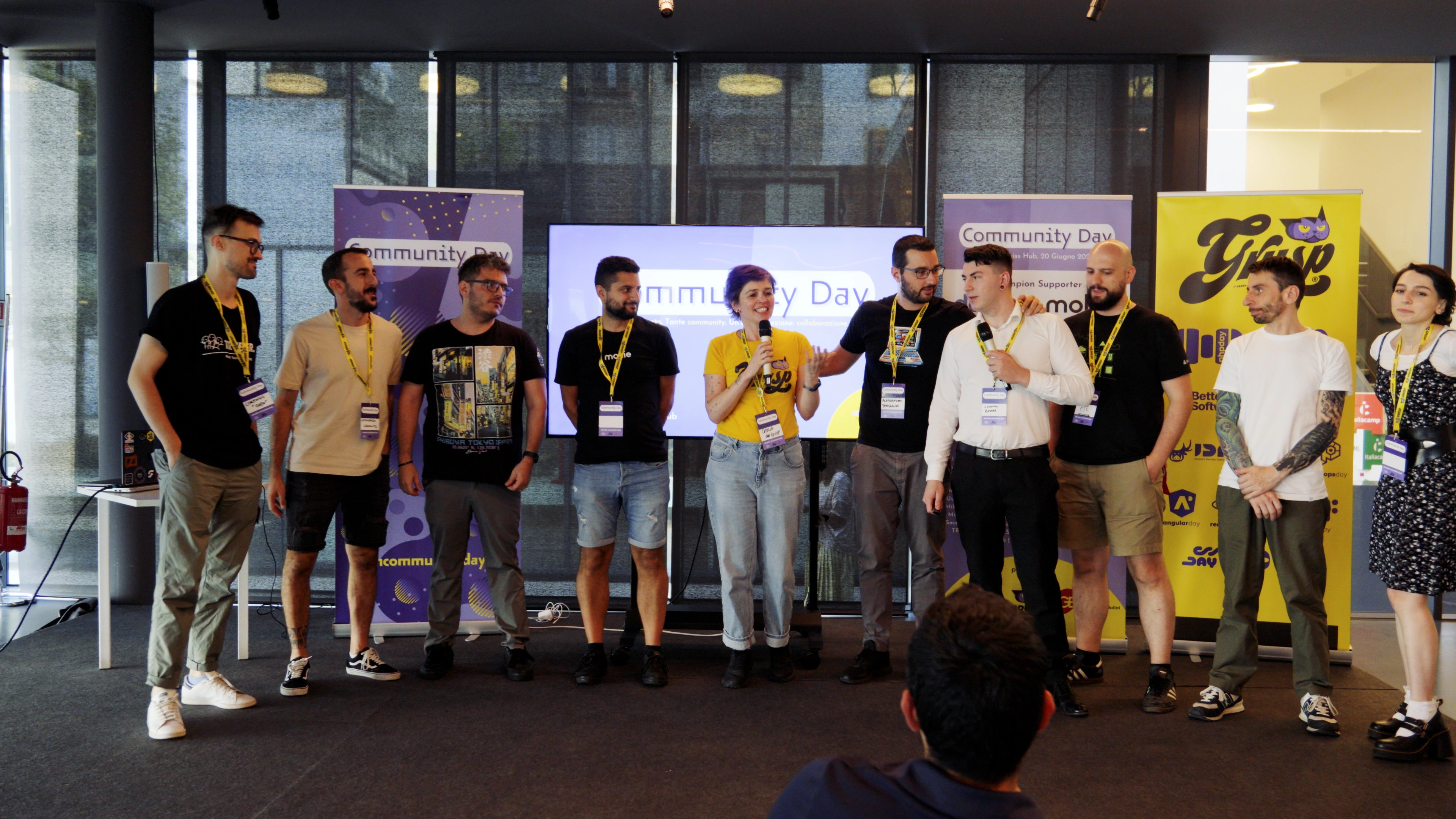 Speakers on stage during the Community Day, with Ludovico Besana among them and event banners in the background.